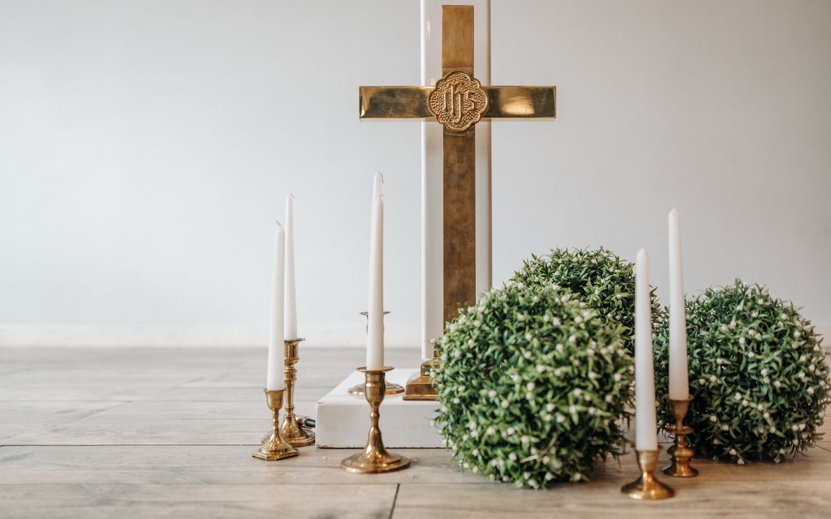 A gold cross with taper candles and greenery on a wooden floor.